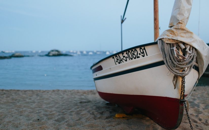 Red and white sailboat sitting on beach with boats in the distance.